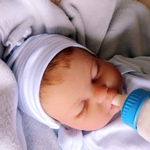 May include: A baby doll wearing a blue and white striped hat and a blue onesie is feeding from a blue and white baby bottle.