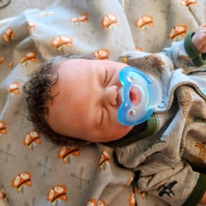 Może przedstawiać: Realistyczna lalka dziecka z brązowymi kręconymi włosami i niebieskim smoczkiem. Lalka ma na sobie szare śpioszki z zielonymi wykończeniami i wzorem zwierząt. Dziecko leży na szarym kocu z pomarańczowymi wzorami lisów.