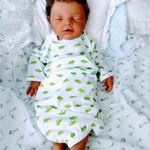 May include: A realistic newborn doll with dark skin and dark hair, wearing a white long-sleeve gown featuring a repeating green dinosaur pattern. The doll is lying on its back with eyes closed, resting on a white fabric surface with a giraffe print.