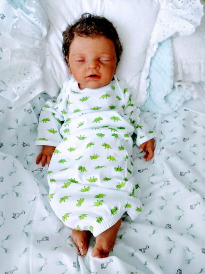 May include: A realistic newborn doll with dark skin and dark hair, wearing a white long-sleeve gown featuring a repeating green dinosaur pattern. The doll is lying on its back with eyes closed, resting on a white fabric surface with a giraffe print.