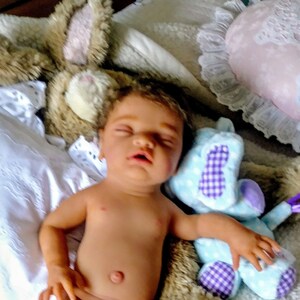 May include: A realistic newborn baby doll with dark hair and closed eyes, lying on a soft surface. The doll is positioned between a tan plush rabbit toy and a light blue plush animal with purple checkered accents, next to white fabric.