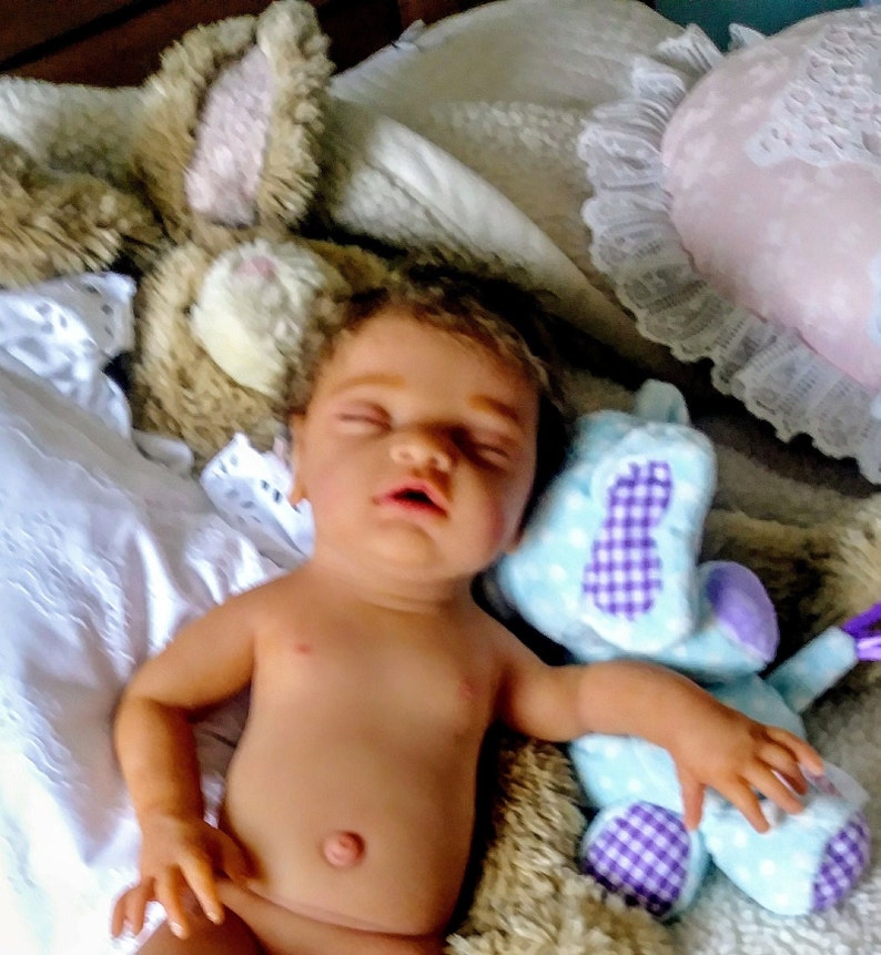 May include: A realistic newborn baby doll with dark hair and closed eyes, lying on a soft surface. The doll is positioned between a tan plush rabbit toy and a light blue plush animal with purple checkered accents, next to white fabric.