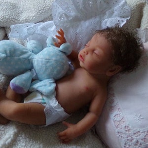 May include: A realistic newborn doll with brown skin and curly brown hair, sleeping while wearing a white diaper. The doll is positioned next to a light blue plush toy with white polka dots and purple ribbon accents, resting on a white textured fabric.