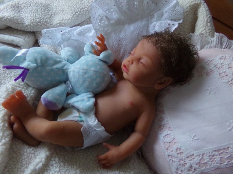 May include: A realistic newborn doll with brown skin and curly brown hair, sleeping while wearing a white diaper. The doll is positioned next to a light blue plush toy with white polka dots and purple ribbon accents, resting on a white textured fabric.
