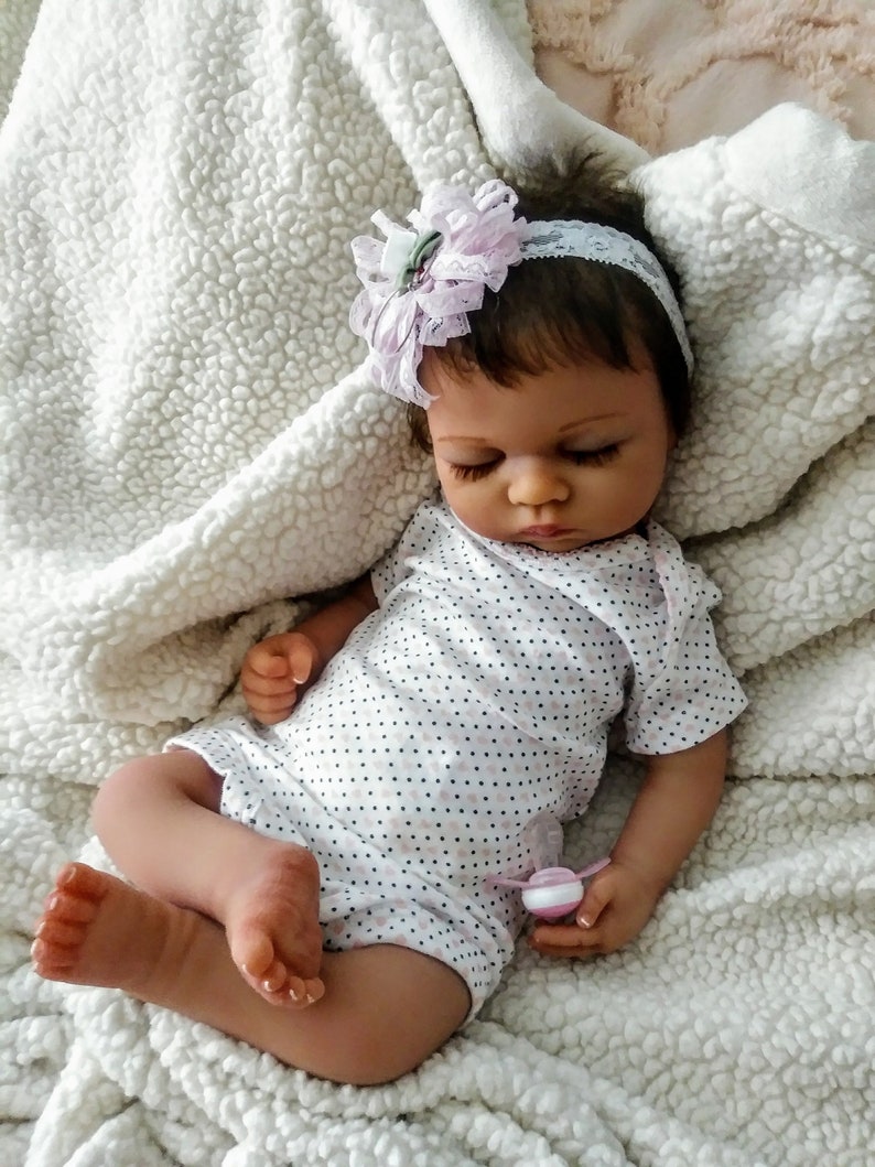 May include: A sleeping baby doll with brown hair wearing a white and black polka dot onesie and a pink flower headband. The doll is lying on a white fluffy blanket.