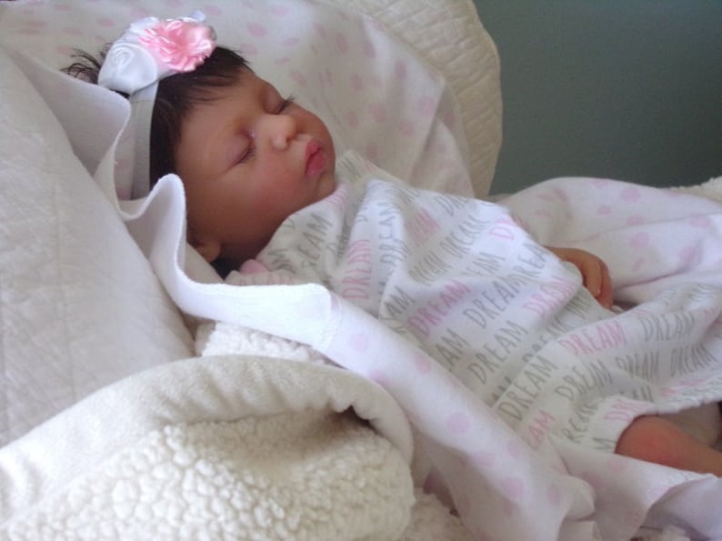 May include: A sleeping baby doll with brown hair wearing a white headband with a pink flower and a white and pink blanket with the text "Dream" repeated.