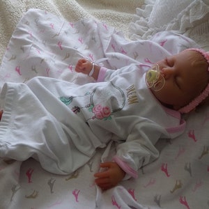 May include: A sleeping reborn baby doll wearing a white and pink long-sleeved gown with a pink headband and a pacifier. The gown has a floral design and the text "Dream" on the front. The doll is lying on a white blanket with a pink and brown pattern.