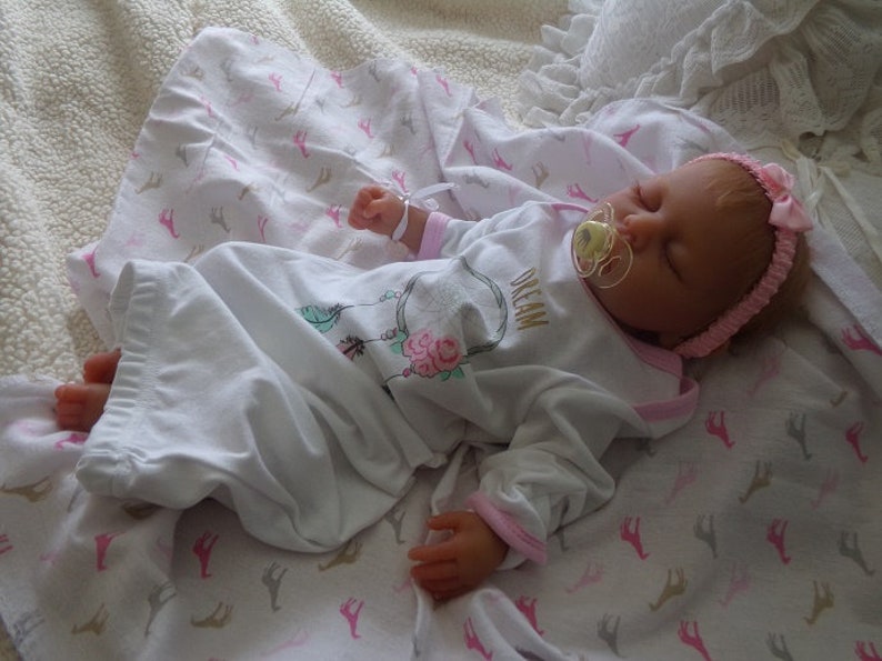 May include: A sleeping reborn baby doll wearing a white and pink long-sleeved gown with a pink headband and a pacifier. The gown has a floral design and the text "Dream" on the front. The doll is lying on a white blanket with a pink and brown pattern.