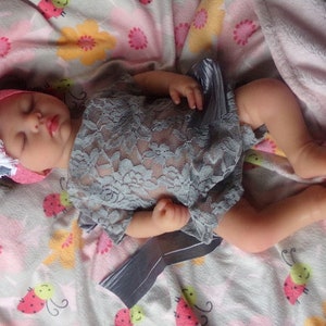 May include: A sleeping baby doll wearing a gray lace dress and a pink headband with a white lace flower accent. The doll is lying on a white and pink blanket with a ladybug pattern.