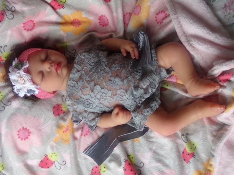 May include: A sleeping baby doll wearing a gray lace dress and a pink headband with a white lace flower accent. The doll is lying on a white and pink blanket with a ladybug pattern.