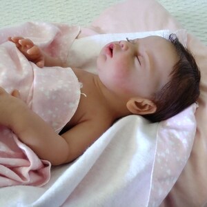 May include: A realistic reborn doll with dark brown hair and closed eyes, lying in a sleeping position. The doll has a light skin tone and is partially covered by a soft, light pink blanket featuring a small white heart pattern throughout the fabric.