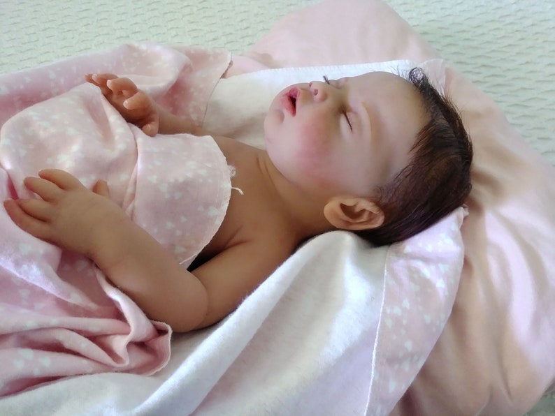 May include: A realistic reborn doll with dark brown hair and closed eyes, lying in a sleeping position. The doll has a light skin tone and is partially covered by a soft, light pink blanket featuring a small white heart pattern throughout the fabric.