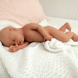May include: A realistic baby doll with light skin tone, closed eyes, and a sleeping expression, lying on its side on a white crocheted blanket. The doll has detailed facial features, small hands, and feet, and is positioned in a resting pose.