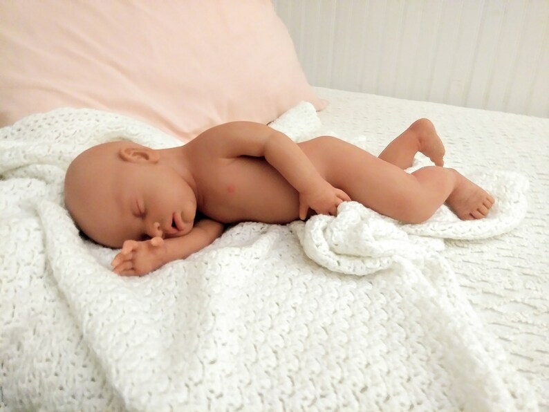 May include: A realistic baby doll with light skin tone, closed eyes, and a sleeping expression, lying on its side on a white crocheted blanket. The doll has detailed facial features, small hands, and feet, and is positioned in a resting pose.
