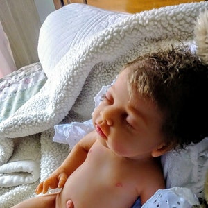 May include: A realistic reborn doll with light skin, closed eyes, and textured dark brown hair. The doll is lying down with its head tilted, featuring detailed facial features, a small nose, and slightly parted lips, resting on a white blanket.