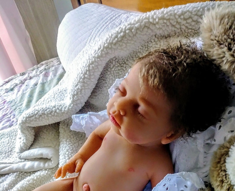 May include: A realistic reborn doll with light skin, closed eyes, and textured dark brown hair. The doll is lying down with its head tilted, featuring detailed facial features, a small nose, and slightly parted lips, resting on a white blanket.