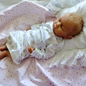 May include: A sleeping baby doll wearing a white floral print long-sleeved onesie, lying on a pink blanket with gold stars, and a white pillow with lace trim.