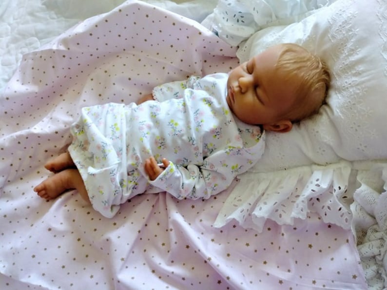 May include: A sleeping baby doll wearing a white floral print long-sleeved onesie, lying on a pink blanket with gold stars, and a white pillow with lace trim.