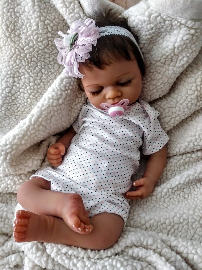 May include: A sleeping reborn baby doll with brown eyes, wearing a white and pink polka dot onesie and a pink and white headband with a flower. The doll is holding a pink pacifier.