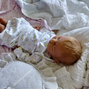 May include: A sleeping reborn baby doll with blonde hair, wearing a white floral print onesie, lying on a white and pink blanket.