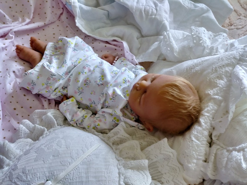 May include: A sleeping reborn baby doll with blonde hair, wearing a white floral print onesie, lying on a white and pink blanket.