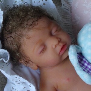 May include: A realistic reborn doll with curly brown hair and closed eyes, sleeping peacefully. The doll has light skin and is resting against a soft white eyelet fabric and a light blue plush toy with a purple and white checkered inner ear detail.