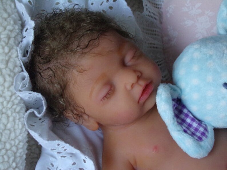May include: A realistic reborn doll with curly brown hair and closed eyes, sleeping peacefully. The doll has light skin and is resting against a soft white eyelet fabric and a light blue plush toy with a purple and white checkered inner ear detail.