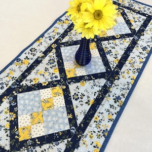 May include: A floral table runner with a blue and yellow color scheme. The runner features a patchwork design with a navy blue border and accents. A blue vase holds yellow flowers, adding a pop of color to the scene. The runner is likely made of fabric.