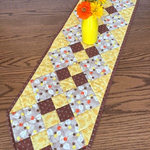May include: A quilted table runner with a patchwork design in shades of yellow, brown, and cream. The runner features a diamond pattern and a brown border. A yellow vase holds orange and yellow flowers.
