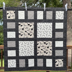 May include: A handmade quilt with a repeating pattern of squares. The quilt features squares with deer and other animal prints, along with solid gray and white squares. The border is black with a paisley design. The quilt is hanging outside.