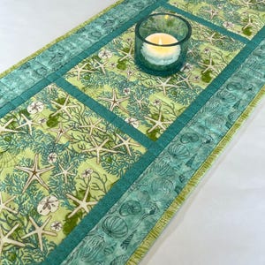 May include: A rectangular quilted table runner featuring a light green fabric with a pattern of white starfish, sand dollars, and coral. The runner has a dark teal border with a seashell print and a teal glass candle holder with a lit candle.