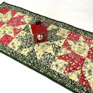 May include: A rectangular quilted table runner featuring a repeating star pattern in red and cream, accented with green fabric printed with pine needles and holly leaves. A small red metal lantern sits on the center of the runner.