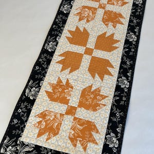 May include: A rectangular table runner featuring a central design of orange-colored bear paw shapes. The runner has a floral patterned background and a black border with white floral accents. The overall design is symmetrical and visually appealing.