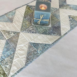 May include: A blue square candle with a lit flame sits on a clear glass holder, tied with a natural raffia bow. The candle rests on a quilted table runner with a geometric pattern in shades of blue, green, and white.
