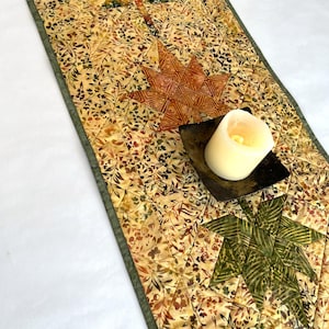 May include: A quilted table runner featuring a beige and brown botanical print with three prominent star-shaped appliqués in dark green, orange, and olive green. A square dark metal dish holds a single white pillar candle on the runner.