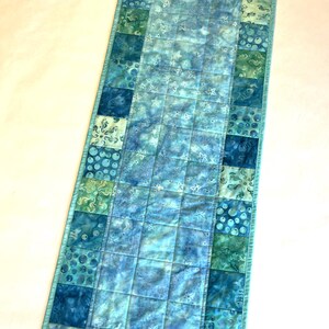 May include: A rectangular quilted table runner featuring a patchwork design in shades of blue and green. The center panel displays a light blue fabric with white starfish motifs, bordered by squares of various aquatic patterns including bubbles and seahorses.