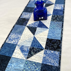 May include: A quilted table runner with a patchwork design in shades of blue and white. The runner features various floral and geometric patterns. A cobalt blue glass vase sits on top of the runner.