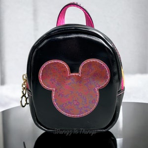 May include: A small black faux leather backpack with a pink zipper and a pink and purple iridescent Mickey Mouse head design on the front. The backpack has a gold chain zipper pull.