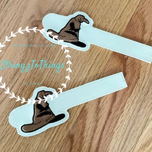 May include: Two light blue fabric bookmarks with a brown embroidered Sorting Hat from the Harry Potter series. The bookmarks are handmade and feature the text "StringzToThings by Diana".