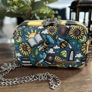 May include: A small, rectangular crossbody bag with a silver chain strap. The bag features a colorful pattern of sunflowers, bees, books, crystals, and clouds on a teal background. The top and sides are yellow.