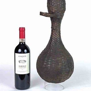 May include: A tall, woven brown decorative vase with a bulbous base and a long neck, featuring a spout-like extension. A bottle of red wine with a red label is next to the vase.