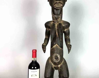 May include: A large, dark wood sculpture of a standing figure with a detailed face and a circular design on the torso. The sculpture is about the height of a wine bottle.