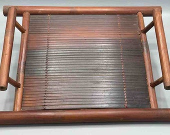 Puede incluir: Una bandeja rectangular de bambú con una superficie de listones y bordes redondeados. La bandeja tiene cuatro soportes verticales y dos soportes horizontales. El bambú es de color marrón cálido.