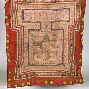 Op de afbeelding: Een rode wandhanger van stof met een geometrisch patroon geborduurd in witte, roze en groene draad. De stof is afgewerkt met een gouden bies en heeft gele pompoenen rond de randen.