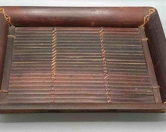 Op de afbeelding: Rechthoekige bamboe dienblad met een donkerbruine afwerking. De lade heeft een lattenbodem en een opstaande rand. De hoeken zijn verstevigd met geweven touw. Een decoratief en functioneel item.