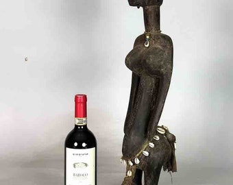 May include: A dark wood sculpture of a woman with a long neck and a beaded necklace. The sculpture is standing next to a bottle of wine.