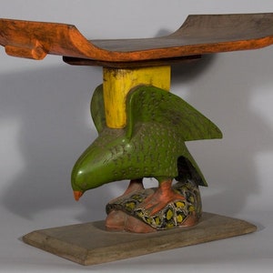 May include: A wooden stool with a flat, curved top and a base shaped like a bird with outstretched wings. The bird is painted green with yellow accents, and the stool is painted orange and brown.