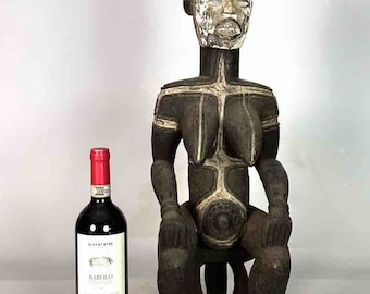 May include: A dark wood sculpture of a seated female figure with white painted details. The figure is wearing a tall, pointed hat and has a circular design on her stomach. The sculpture is standing on a white surface next to a bottle of wine.