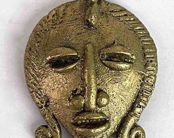 Puede incluir: Un colgante de metal dorado en forma de rostro humano estilizado. El rostro tiene los ojos cerrados, una nariz prominente y una boca ancha. El colgante tiene un bucle en la parte superior para colgar.