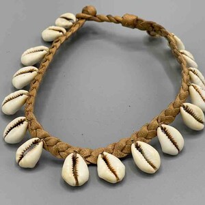Op de afbeelding: Een bruin leren koordarmband met witte kauri schelpen eraan bevestigd. De armband is gevlochten en heeft een eenvoudig ontwerp.